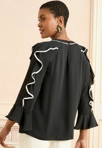 Blusa nera con dettagli in pizzo bianco lungo le maniche e il colletto. Presenta maniche lunghe e un'apertura a forma di chiave sul retro. Tessuto di consistenza liscia.