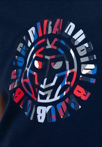 Camiseta azul marino con un gráfico circular que presenta un rostro estilizado en rojo, azul y gris. Elementos de texto dispuestos alrededor del diseño.