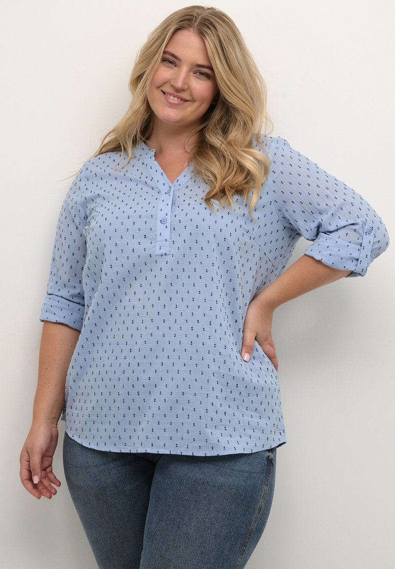 Camisa azul claro con tapeta de botones, mangas de tres cuartos y un patrón texturizado de pequeños puntos azul oscuro en todo su diseño.