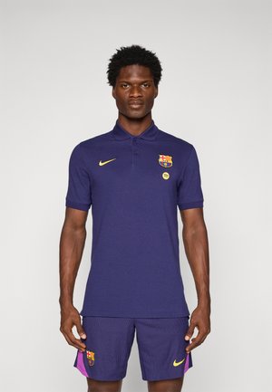 Mand står iført en mørkeblå FC Barcelona polo shirt og matchende shorts med gule Nike-logoer og klublogoer.
