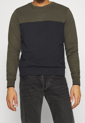 Hombre con sudadera de manga larga en bloque de colores verde oliva y negro, y vaqueros gris oscuro, de pie contra un fondo claro y liso.