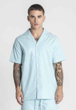 Junger Mann mit kurzen Haaren und Arm-Tattoos, der ein hellblaues kurzärmeliges Hemd mit Knöpfen und passende Shorts vor einem schlichten Hintergrund trägt.