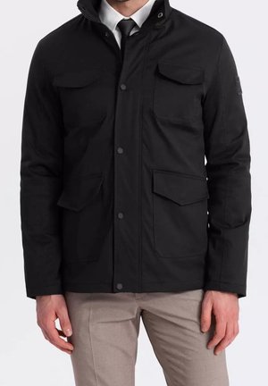 Homme portant une veste noire avec quatre poches à rabat sur le devant, une chemise blanche, une cravate noire et un pantalon beige sur un fond uni.