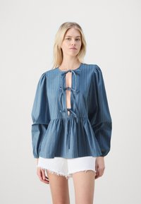 Denima blūze ar vertikālām baltām svītrām, apaļu kakla izgriezumu un priekšpusi ar atvēruma formu, kas piestiprināta ar saitēm. Puffētas piedurknes un pepluma viduklis. Baltas plānās šortus.