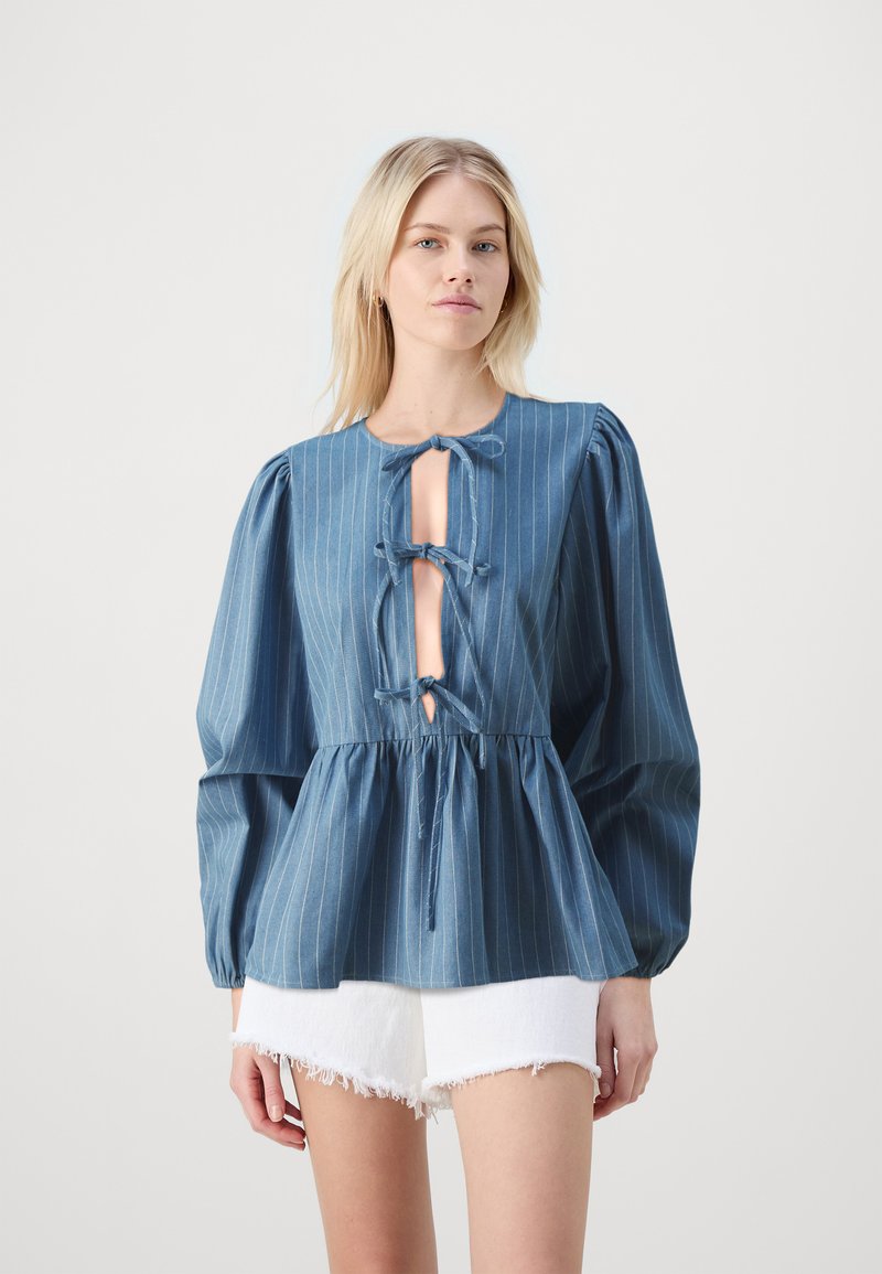 Denima blūze ar vertikālām baltām svītrām, apaļu kakla izgriezumu un priekšpusi ar atvēruma formu, kas piestiprināta ar saitēm. Puffētas piedurknes un pepluma viduklis. Baltas plānās šortus.