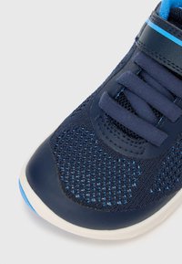 Marineblå sportsko med lufthull øvre del, polstret ankelklaff og syntetiske detaljer. Har flate lisser og en kontrasterende lyseblå stripe.