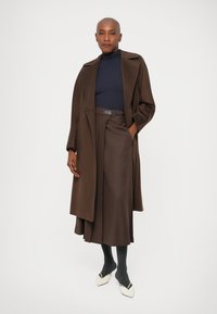 Mulher careca com um longo casaco castanho, saia plissada combinando, suéter preto, meias escuras e sapatos brancos de bico fino, de pé com uma mão no bolso.