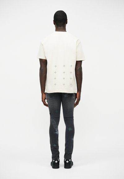 Homme debout de dos à la caméra, portant un t-shirt blanc à signes plus gris, un jean noir avec des taches délavées et des chaussures noires sur fond blanc.