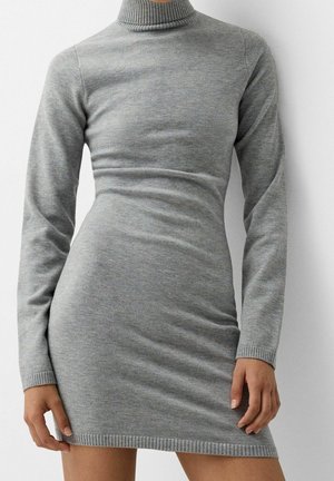 Figursyet lysegrå langærmet rullekravesweaterkjole med ribstrikkede manchetter og kant, båret af en person, der står mod en ensfarvet baggrund.