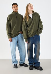Olive zelena zip-up dukserice, ležeran kroj, uparene sa svijetloplavim i gradijentno tamnoplave široke traperice. Oba modela nose crne cipele.