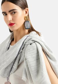 Sciarpa grigia chiara e tessurizzata appoggiata sulla spalla, caratterizzata da linee orizzontali. Orecchini geometrici di grandi dimensioni con un mix di materiali dorati e neri.