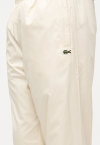 Μπεζ casual παντελόνι με ελαστική μέση, πλαϊνή τσέπη και μικρό πράσινο κεντημένο λογότυπο κροκοδείλου στο δεξί μηρό.