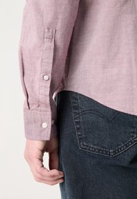 Gros plan sur le bras gauche d'une personne portant une chemise rose à manches longues et un jean en denim bleu foncé, la main détendue le long du corps.