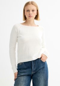Jersey blanco de punto con un cuello ancho, mangas largas y dobladillo acanalado, combinado con unos vaqueros azul de talle alto.