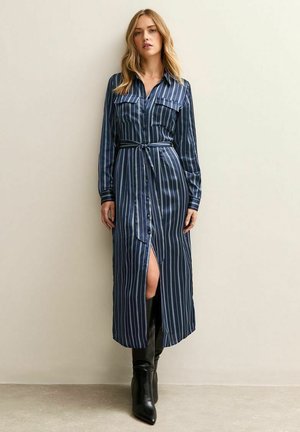 Donna che indossa un lungo abito a righe blu navy con bottoni e cintura in vita, abbinato a stivali neri fino al ginocchio, in piedi contro un muro neutro.