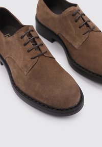 Zapatos de ante marrón con cordones y punta redonda, suela de goma oscura y detalles de costura en contraste. Diseño clásico con acentos mínimos.