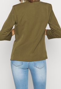 Olive green blazer with structured shoulders, 3/4 ruched sleeves, and a fitted design. Worn with light blue denim jeans.