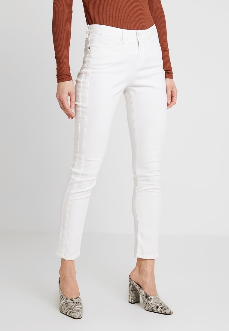 Femme portant un jean blanc slim, un haut marron à manches longues côtelé et des mules à talons hauts imprimé serpent, debout sur un sol gris clair.
