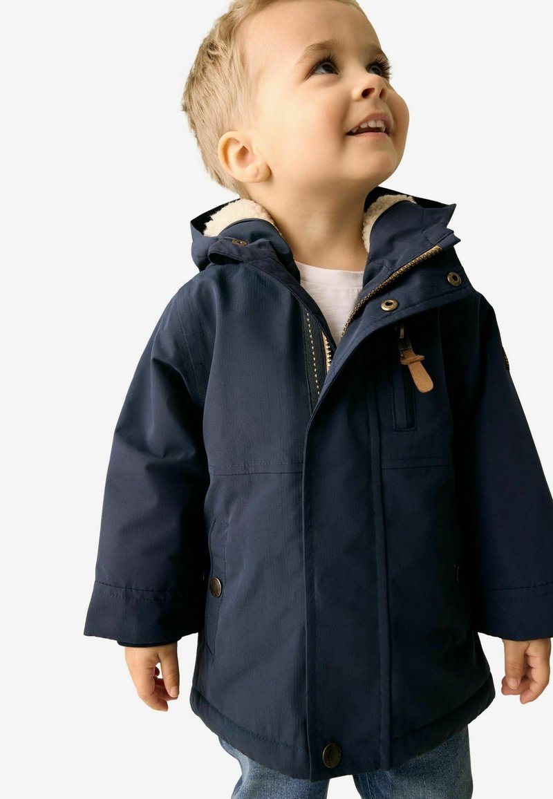 Veste isolante bleu marine avec une capuche en fourrure, fermeture éclair à l'avant, boutons-pression et poches latérales. Le tissu est lisse avec une finition brillante.