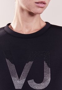Sweatshirt noir avec un col rond présentant un graphique de strass dégradé des lettres "VJ" sur le devant. Texture lisse.