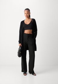 Top court noir, cardigan long et pantalons larges. Tissu lisse avec une coupe décontractée. Sac à main noir et sandales à glissière pour un look complet.