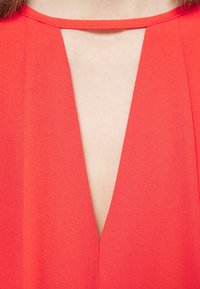 Gros plan d'une personne portant un vêtement rouge avec un décolleté en V profond et une lanière horizontale étroite sur la poitrine.