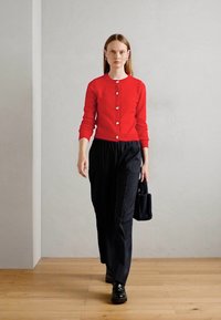 Červený pletený cardigan s kulatým výstřihem a stříbrnými knoflíky, kombinovaný s černými pruhovanými kalhotami a malou černou kabelkou.
