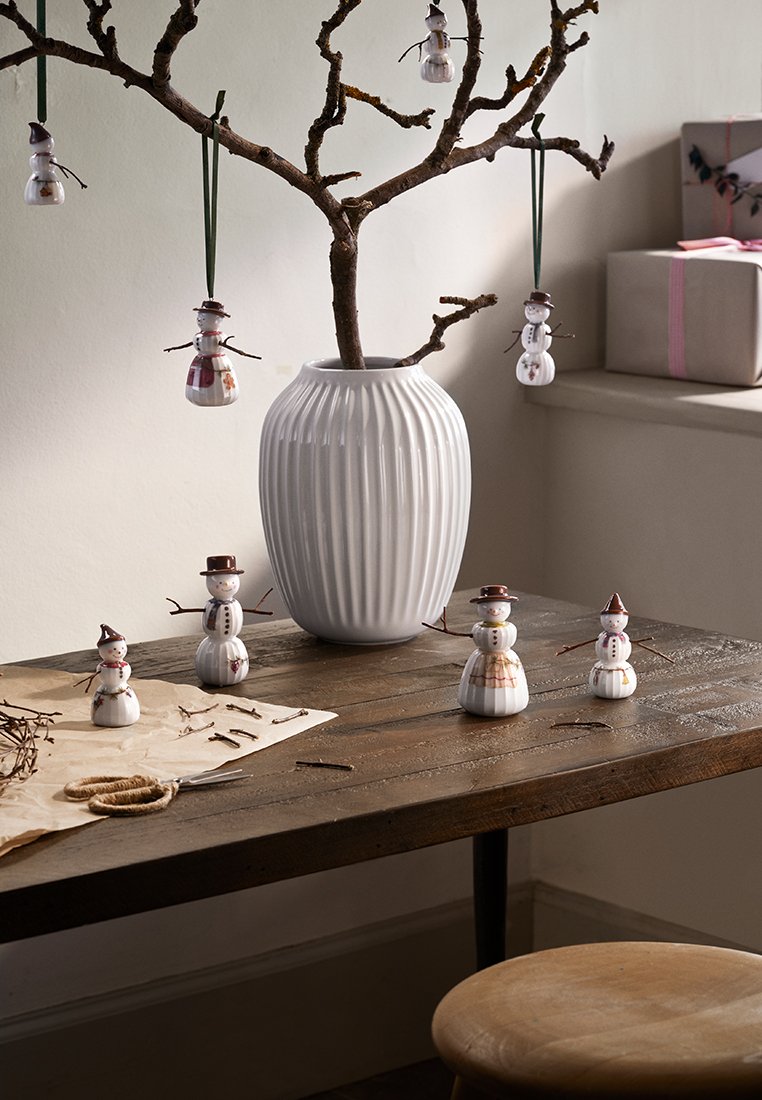 Keramische Schneemänner mit braunen Hüten stehen auf einem Holztisch. Eine gerippte weiße Vase hält einen kahlen Zweig, der mit hängenden Schneemann-Ornamenten geschmückt ist.