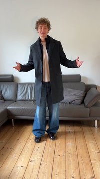 Eine Person steht in einem grauen Mantel über einem weißen Hemd, kombiniert mit weitem blauen Jeansstoff und schwarzen Schuhen, in einem modernen Raum mit einem Sofa.