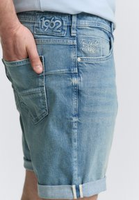 Jasnoniebieskie szorty dżinsowe z podwiniętymi nogawkami, z tylną kieszenią, haftowanym logo i wyblakłą teksturą. Szczegółowe przeszycia na całej powierzchni.