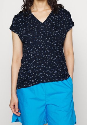 Kvinde iført en sort V-hals-bluse med små blå blomsterprint og lyseblå shorts, stående med den ene hånd i lommen.