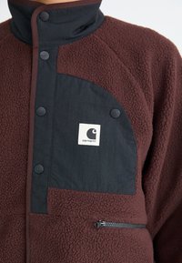 Chaqueta de forro polar marrón con cuello abotonado negro y bolsillo en el pecho que presenta un parche con el logo blanco de Carhartt y un bolsillo lateral con cremallera.