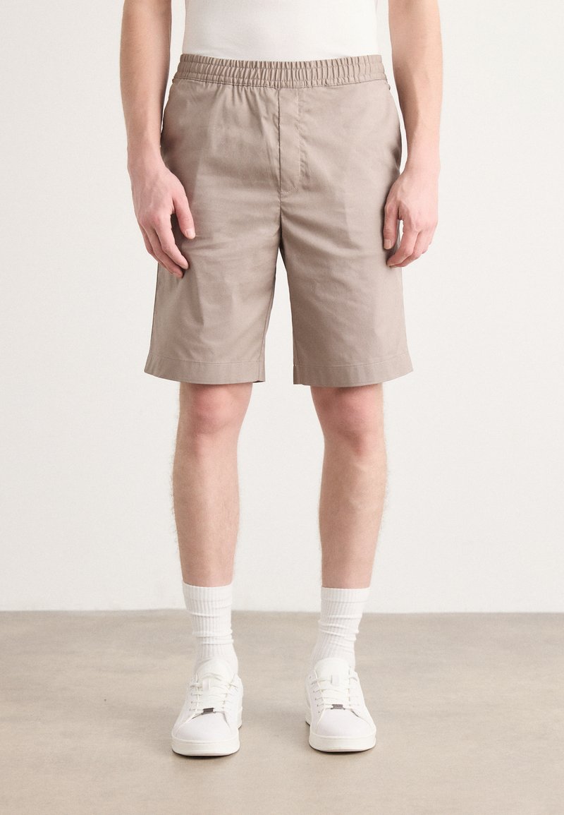 Beige shorts med elastisk talje, knæ-længde snit og glat tekstur, iført hvide sokker og sneakers på en neutral baggrund.