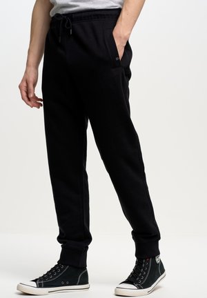 Pantalones de chándal negros con cintura elástica ajustable, bolsillos laterales y puños acanalados, combinados con zapatillas altas negras. Textura suave.