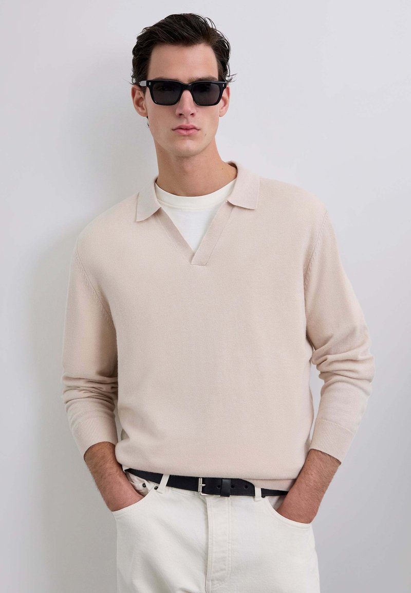 Pull en maille beige clair avec un col en V et un col rabattu, associé à un t-shirt blanc. Porté avec un pantalon crème et des lunettes de soleil noires.