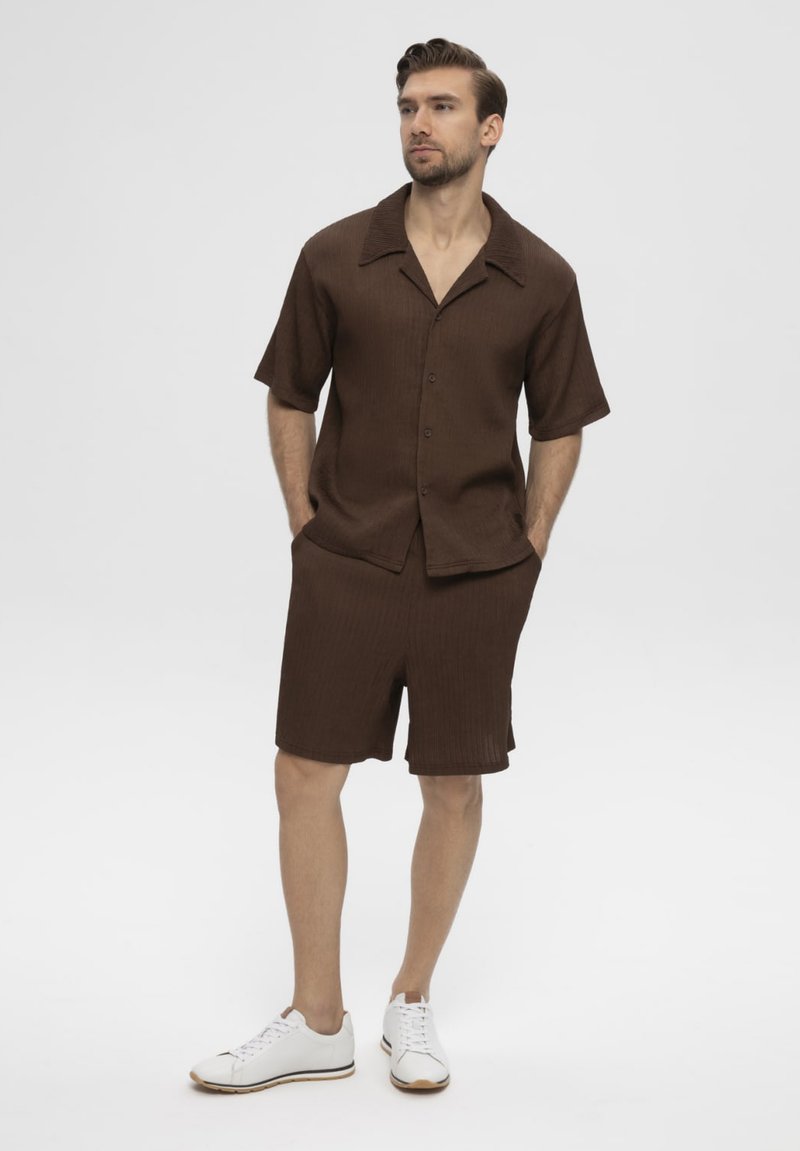 Homme debout les mains dans les poches, portant une chemise marron à manches courtes boutonnée, un short assorti et des baskets blanches sur fond uni.