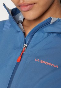 Chaqueta azul con capucha con cremallera, que presenta un forro gris en contraste, un logotipo prominente y un tirador de cremallera naranja. Tejido suave y ligero.