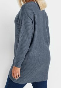 Jersey de punto gris con mangas largas, un corte holgado y patrón texturizado. La prenda llega por debajo de las caderas, destacando la comodidad.