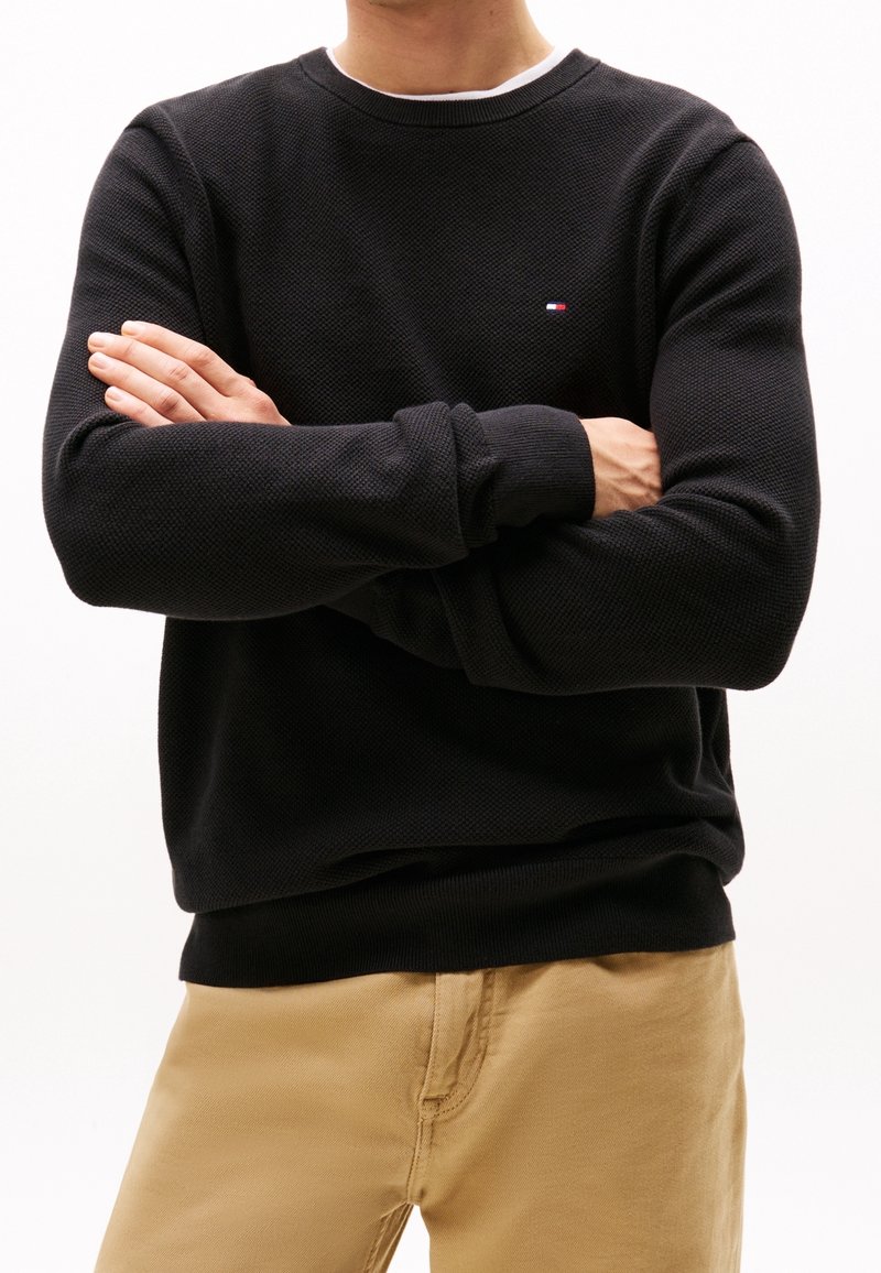 Personne portant un pull texturé noir avec un petit logo sur la poitrine et un pantalon beige, bras croisés devant un fond blanc uni.