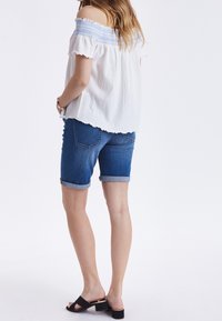 Top bianco off-shoulder con dettagli ricamati blu, abbinato a pantaloni corti di jeans arrotolati. Sandali neri con tacco a blocco completano il look.