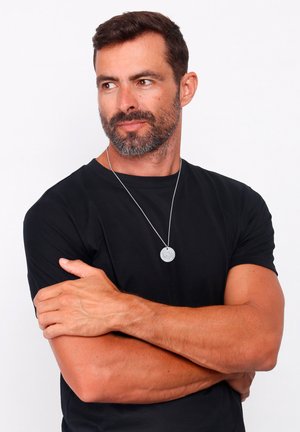 Ciondolo circolare in argento sterling su una catena sottile, indossato sopra una t-shirt nera. Il soggetto ha le braccia incrociate, una carnagione chiara e capelli corti marroni.