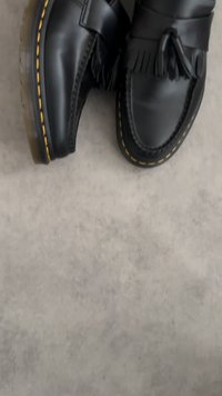 Juodos odinės su kutais loafer tipo bateliai su geltonomis siūlėmis ant permatomų guminių padų, ilsintis ant pilkos tekstūruotos paviršiaus.
