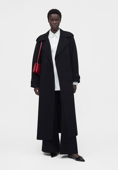 Μαύρη γυναίκα που φορά ένα μακρύ μαύρο παλτό με διπλό πέτο, λευκό πουκάμισο με κουμπιά, φαρδιά μαύρα παντελόνια, μαύρα μυτερά παπούτσια και κρατά μια κόκκινη τσάντα ώμου.