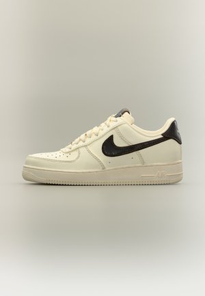 Μπεζ χαμηλά sneakers Nike Air Force 1 με μαύρο λογότυπο swoosh, μπεζ κορδόνια και μπεζ σόλα σε ουδέτερο φόντο.