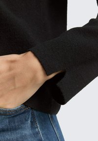 Zwarte gebreide trui met een gespleten manchet detail, dat een zachte textuur vertoont. De hand steunt op spijkerbroek met zichtbare stiksels.