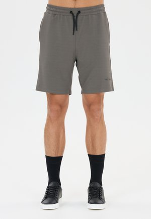 Mand iført grå snøre-shorts, sorte mellemlange sokker og sorte lave sneakers med hvide såler, stående mod en ensfarvet baggrund.