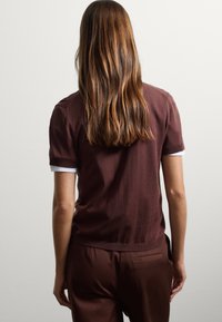 Top de punto de algodón marrón con cuello acentuado en blanco, mangas cortas y un ajuste relajado. La modelo aparece de espaldas, con el cabello cayendo sobre los hombros.