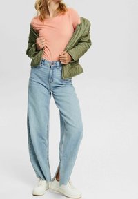 Grüne Pufferjacke über einem pinken T-Shirt, kombiniert mit weiten, hellblauen Jeans mit seitlichen Schlitzen und weißen Sneakers.