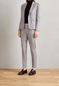 Szary garnitur w kratę z dopasowaną marynarką i zwężanymi spodniami. Charakteryzuje się teksturowaną tkaniną, złotymi guzikami oraz noszony jest z ciemnymi mokasynami.