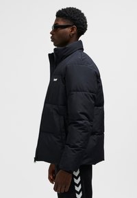 Sort dunjakke med høj krave, lynlåslukning og sidelommer. Glat tekstur med vandrette quiltede sektioner og et logo-emblem.